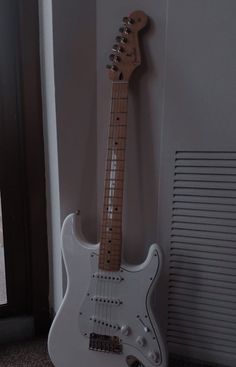 a white electric guitar sitting on the floor next to a wall with a window behind it