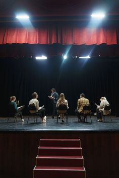 five people sitting on chairs in front of a stage with red curtains and steps leading up to them