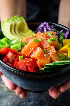 a person holding a bowl filled with assorted veggies and sliced avocado