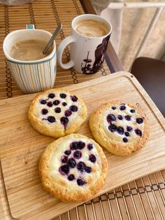 three blueberry cookies sitting on top of a wooden cutting board next to two cups of coffee