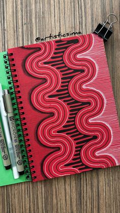 a red notebook with black and white swirls on it next to two markers,