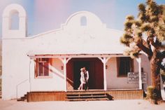 a man and woman standing on the porch of a small white building with palm trees