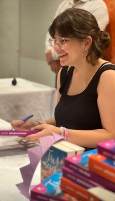 a woman is sitting at a table with books and writing on her notebook while smiling