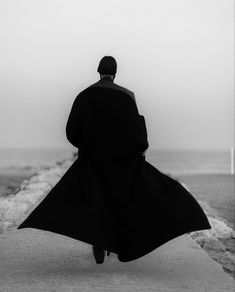 a man in a long black coat is walking on the sidewalk by the ocean with his back to the camera