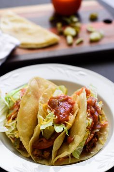 three tacos on a white plate with lettuce and tomato in the background