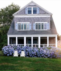 a house with blue flowers in front of it and two lawn chairs on the grass