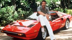 a man sitting on the hood of a red sports car in front of some trees