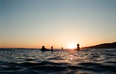 two people are swimming in the ocean at sunset or dawn, with one person sitting on his surfboard