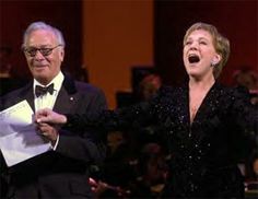 an older man and woman singing together in front of a crowd with their arms outstretched