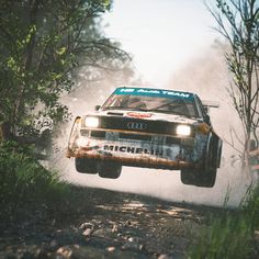 an old race car is making a jump in the air on a dirt road with trees and bushes behind it