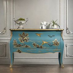 a blue dresser with gold trim and flowers on the top, in front of silver wall paneling