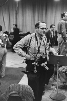 black and white photograph of man standing in front of microphones with other people around him