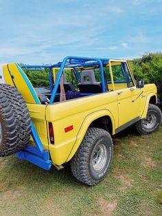 a yellow truck with large tires parked on the grass in front of some bushes and trees