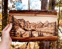 a person holding up a piece of wood with mountains and trees on it in the woods