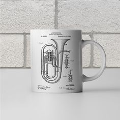 a coffee mug with an illustration of a french horn on the front and side, sitting against a white brick wall