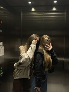 two women standing in front of an elevator taking pictures with their cell phones and looking into the mirror