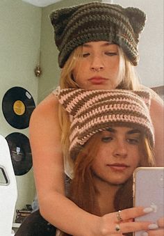 two women wearing knitted hats while looking at a cell phone in a room with records on the wall