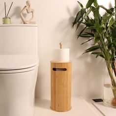 a white toilet sitting next to a plant and a wooden trash can on top of a counter