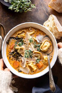 two hands holding a bowl of soup with tortellini and carrots in it