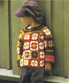 a little boy standing next to a green door wearing a red and yellow sweater with flowers on it