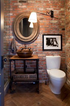 a bathroom with a toilet, sink and mirror on the brick wall above it is a wood floor