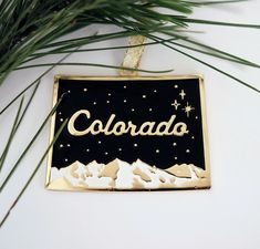 a black and gold christmas ornament with the word colorado on it hanging from a pine tree