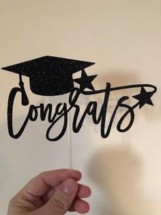 a hand holding up a cake topper that says congrats with a graduation cap on it