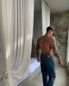 a man with tattoos standing in front of a curtained window looking at the bed