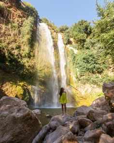 a woman standing at the base of a waterfall with her back turned to the camera