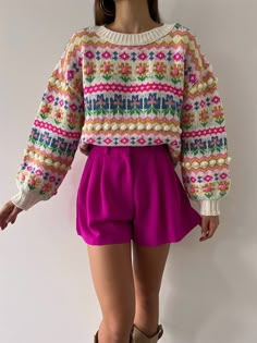 a woman is standing in front of a white wall wearing shorts and a colorful sweater