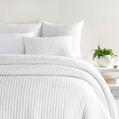 a bed with white comforters and pillows in a room next to a plant on a table