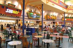 the inside of a restaurant with tables, chairs and people walking around in front of it