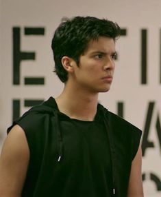 a young man standing in front of a white wall wearing a black hoodie and looking off to the side