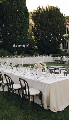 a long table set up with white linens and place settings for an outdoor dinner