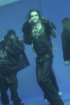 a man with long hair standing in front of a blue background wearing black leather clothes