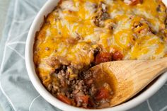 a casserole dish with meat, cheese and tomatoes in it next to a wooden spoon