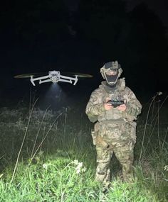 a man in camouflage holding a camera next to a remote control helicopter on the grass