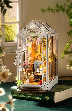 a miniature house on top of a book with flowers in the window sill behind it