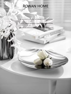 white flowers are sitting on a glass plate next to a book and vase with books in the background