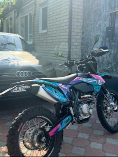 a pink and blue dirt bike parked in front of a building next to a car