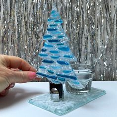 a hand is holding a small glass christmas tree on a table with silver sequins behind it