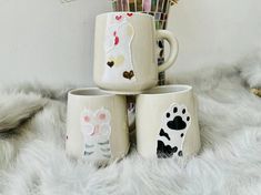 three coffee mugs with designs on them sitting on a fur rug next to pens and pencils