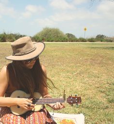 a woman sitting in the grass playing an ukulele with a hat on her head