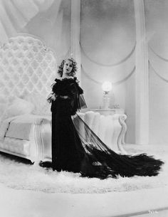 an old fashion photo of a woman in a black dress standing next to a bed