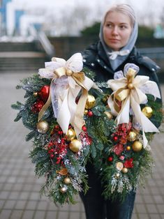 a woman holding two wreaths with gold and white bows on them in front of a brick walkway