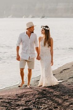 a man and woman holding hands walking on the beach next to the water with boats in the background