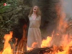 a woman standing in front of a fire with her hands up to the sky and mouth open