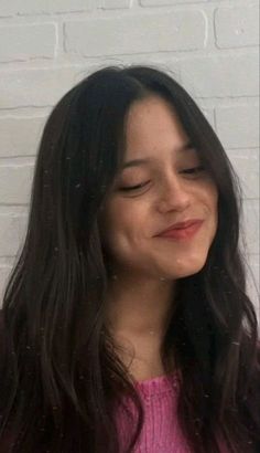 a woman with long black hair smiling and holding a cell phone to her ear while standing in front of a white brick wall