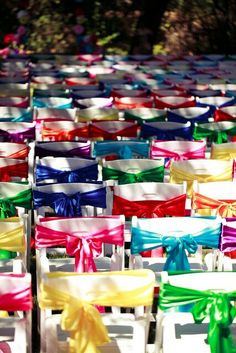 rows of colorful chairs with bows tied to them in front of trees and bushes, text reads rainbow wedding