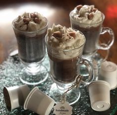 three cups filled with hot chocolate on top of a glass table topped with whipped cream and nuts